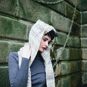 May include: A woman wearing a gray hooded sweater with a white and brown knit scarf. She is standing in front of a brick wall with a chain link fence.
