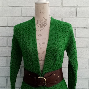 May include: A green crocheted cardigan with a brown leather belt. The cardigan has a V-neck and long sleeves. The belt has a gold buckle and is fastened at the waist.