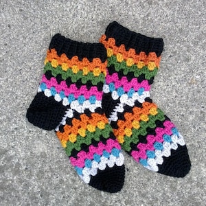 Puede incluir: Un par de calcetines de ganchillo coloridos. Los calcetines tienen un puño, talón y puntera negros, con un patrón vibrante de cuadrados naranjas, verdes, rosas, azules y blancos. Los calcetines están hechos a mano.