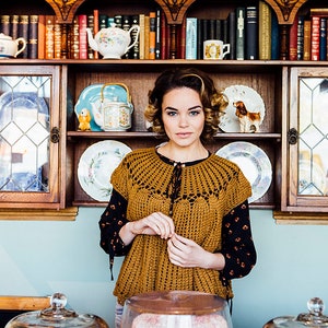 Könnte beinhalten: Eine Frau in einem braunen Häkeltop steht vor einer Vitrine mit Kuchen unter Glaskuppeln. Die Vitrine befindet sich hinter einer Theke aus Holz mit einem Regal darüber. Das Regal ist gefüllt mit Teetassen, Teekannen und Büchern.