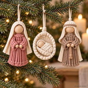 May include: Three macrame Christmas ornaments featuring two figures with wooden bead heads and cream-colored head coverings, one in a dusty rose dress and one in a tan dress, alongside a central circular macrame cradle with a swaddled infant.