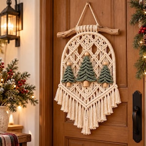 May include: A macrame wall hanging featuring a cream-colored knotted cord design suspended from a natural wooden branch. The piece includes three green cord-knotted pine tree shapes with wooden bead trunks and a long, fringed bottom edge.