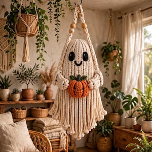 May include: A macrame ghost wall hanging made of cream-colored cord, featuring black embroidered eyes and a smiling mouth. The ghost figure holds a small, textured orange pumpkin with a green stem in its center and hangs from a braided cord.