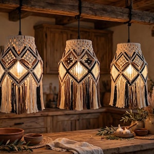 Puede incluir: Tres lámparas colgantes con pantallas de macramé intrincadas en cordón color crema, marrón y negro. Cada pantalla muestra un patrón geométrico de diamantes con flecos largos y superpuestos en la base, suspendidas de una viga de madera.