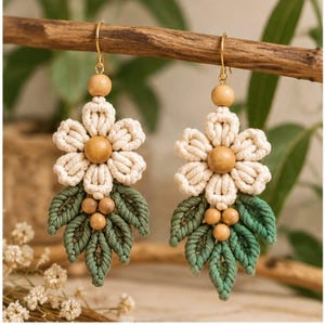 Puede incluir: Un par de pendientes de macramé con pétalos de cordón color crema en forma de flor y una cuenta de madera central. Debajo de la flor, tres hojas de macramé verde están acentuadas por tres pequeñas cuentas de madera en ganchos dorados.