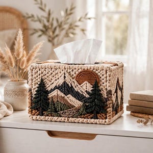 May include: A rectangular tissue box cover crafted from cream-colored cord, featuring a detailed macrame landscape design. The scene depicts dark green pine trees, layered mountain peaks in shades of brown and cream, and a circular sun in rust.