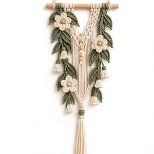 May include: A macrame wall hanging suspended from a wooden dowel, featuring cream-colored cord, olive green leaf accents, and three cream flowers with wooden bead centers. The design includes vertical cord patterns and a long bottom tassel.