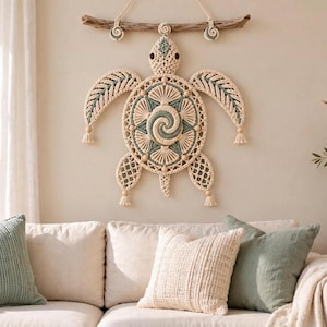 Puede incluir: Un tapiz de pared con una tortuga marina de macramé hecha de cuerda color crema y verde salvia. La tortuga está suspendida de un trozo de madera flotante y muestra intrincados patrones de nudos, diseños de conchas y pequeñas borlas.