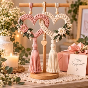 Puede incluir: Dos adornos de macramé en forma de corazón colgados de un soporte de madera. Un corazón es rosa empolvado y el otro es crema, ambos con flores de ganchillo, hojas verdes y borlas largas. Un pequeño corazón de madera cuelga entre ellos.