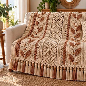 May include: A cream and terracotta colored macrame throw blanket featuring intricate vertical patterns of raised leaf motifs, diamond shapes, and textured knots. The blanket is finished with a long fringe of alternating cream and terracotta tassels.