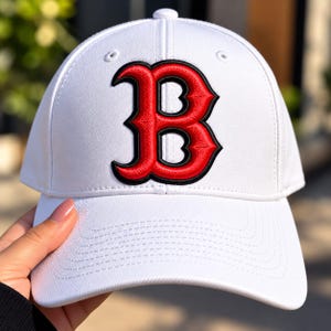 May include: A white baseball cap featuring a large, embroidered letter B on the front. The letter is red with a thick black outline, rendered in a stylized, gothic-inspired font. The cap has a structured crown, a curved brim, and eyelets.