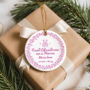 May include: A round white ceramic ornament with a gold string, featuring a pink floral wreath design, a teddy bear icon, and the text First Christmas as a Mama, Olivia Rose, 10.14.2026, 7 lb 2 oz printed in pink ink.