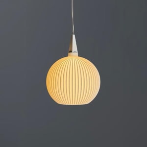 Könnte beinhalten: Eine kugelförmige Pendelleuchte mit einem gerippten, cremeweißen Keramikschirm, der ein warmes Licht ausstrahlt. Die Lampe hängt an einem dünnen weißen Kabel, das an einer silberfarbenen, konischen Metallkappe oben am runden Schirm befestigt ist.