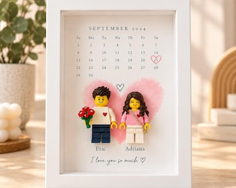 May include: A white framed wall art piece featuring a September 2024 calendar with the 21st circled in red. Below, two plastic toy figures stand before a pink watercolor heart. The names Eric and Adriana are printed with the text I love you so much.