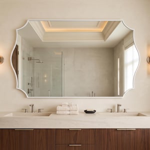 May include: A wall-mounted mirror with a white frame and a scalloped edge, positioned above a double vanity with two sinks, a light-colored countertop, and dark wood cabinetry. Two vertical wall sconces flank the mirror on either side.