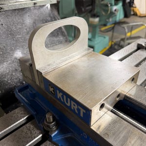 May include: A silver metal Kurt machine vise jaw with a vertical handle featuring an oval cutout. The jaw has a blue side panel with the white text KURT printed on it. The component is mounted on a blue metal base with a threaded bolt.
