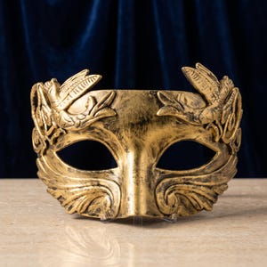 May include: A gold-colored masquerade mask with a distressed finish, featuring sculpted bird motifs on the upper corners and intricate leaf-like scrollwork along the lower edges, designed to cover the eye area with two large openings.