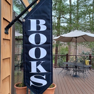 May include: A black banner with the white word "BOOKS" vertically displayed. The banner is hanging outside a building. The background includes a deck with outdoor furniture and a beige umbrella.