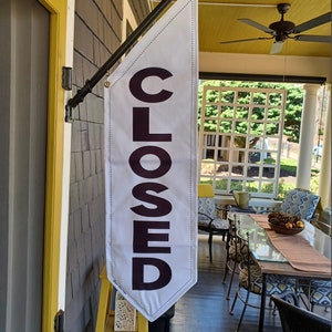 May include: A white flag with black text that reads "CLOSED" hanging from a black metal pole. The flag is attached to the side of a building with a yellow door.