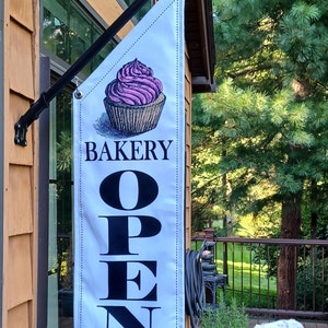 May include: White bakery sign with a cupcake illustration and the word "BAKERY" above the word "OPEN" in large black letters. The sign is hanging outside a building with a brown exterior. A white dog is resting on a wooden deck.