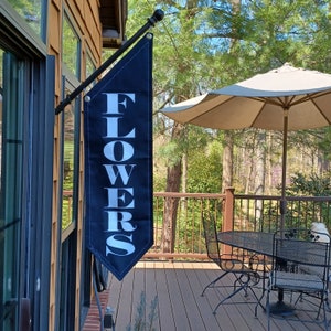 May include: A black flag with white lettering that reads "FLOWERS" hanging from a black metal pole on a wooden porch. The flag is hanging in front of a window with a view of a wooded area. A white patio umbrella is in the background.