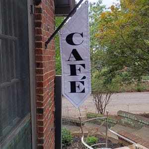 May include: A white cafe flag with black text hangs from a black metal rod attached to a brick building. The flag reads "CAFÉ" in a bold, serif font. The scene includes a door, a chair, and greenery.
