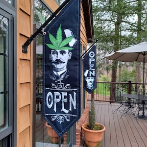 May include: Black and green open sign with a cartoon image of a man with a leafy green hat and the word "OPEN" in white lettering.