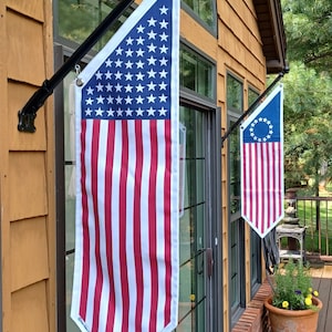 May include: Two American flags hanging from a black metal flagpole outside a wooden house. The flag on the left is the standard American flag, while the flag on the right is the Betsy Ross flag.