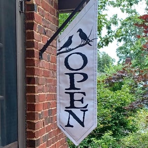 May include: White rectangular hanging sign with the word "OPEN" in large black letters. Two black bird silhouettes perch on a branch above the text. The sign is mounted on a black metal bracket attached to a brick wall.