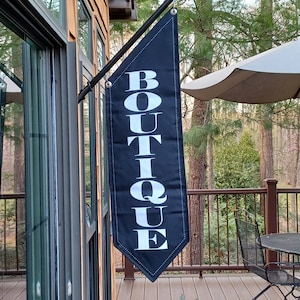 May include: A black boutique flag with white text hangs outside. The flag is rectangular with a pointed bottom and is attached to a black pole. The word "BOUTIQUE" is vertically displayed in large, bold letters.