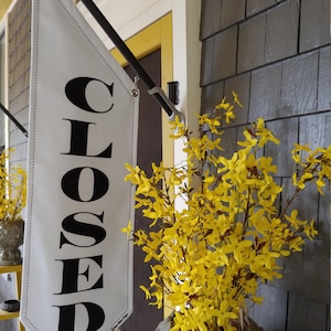 May include: A white banner with black text that reads "CLOSED" hanging from a black metal pole. The banner is attached to a building with yellow siding. A bouquet of yellow flowers is in a gray pot in the foreground.