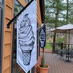 May include: Two black and white ice cream cone flags hanging on a wooden building. The ice cream cones are drawn in a vintage style. The flags are hanging from a metal pole.