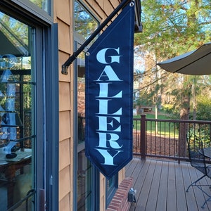 May include: A navy blue outdoor banner with the word "GALERY" in white capital letters. The banner is hanging from a black metal pole attached to a house with brown siding. The banner is rectangular with a pointed bottom.
