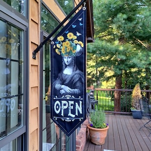 May include: A black banner with the Mona Lisa wearing a sunflower headdress and butterflies. The banner reads "OPEN" in white script. The banner is hanging outside a building with a wooden exterior.