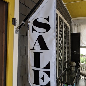May include: A white banner with black lettering that reads "SALE" hanging from a black pole. The banner is attached to a building with a yellow door.