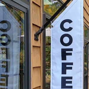May include: A white banner with black text that reads "COFFEE" hanging from a black metal flagpole outside a building with a wooden exterior and large windows.