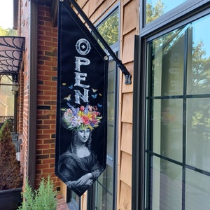 May include: Black and white sign with the word "OPEN" in white letters. The sign is decorated with a floral wreath and a woman's face. The sign is hanging from a black metal pole.