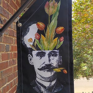 May include: A vertical black garden flag featuring a black and white illustration of a man with a mustache holding a tulip in his mouth. The top of the man's head is open, revealing a bouquet of orange, yellow, and pink tulips with green leaves.