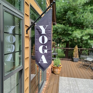 May include: A black yoga sign with white lettering hanging on a wooden building. The sign is attached to a black metal pole. The sign is hanging outside a building with a wooden deck.