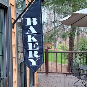 May include: A black banner with the word "BAKERY" in large white letters hangs outside a building. The banner is mounted on a black metal arm. The building has brown siding and windows. A deck with a table and chairs is visible.
