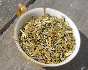 May include: A white ceramic cup with a gold rim filled with dried green herbs. The cup is sitting on a wooden surface.