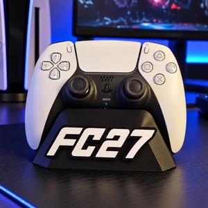 May include: A white and black wireless video game controller resting on a black plastic display stand. The stand features the white text FC27 embossed on the front. The controller has two analog sticks, a directional pad, and four action buttons.