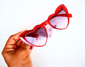 May include: Red heart-shaped sunglasses with a pink tint. The sunglasses are held in a hand.