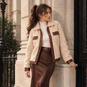 May include: A cream-colored sherpa jacket featuring brown faux leather trim, a pointed collar, two chest pockets with snap buttons, and a front button closure. The jacket is worn over a white turtleneck sweater and a brown midi skirt.