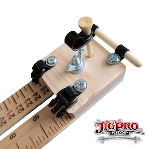 May include: A wooden jig with a ruler, two black clamps, and a golf tee with a wooden dowel attached. The jig is used for woodworking projects.