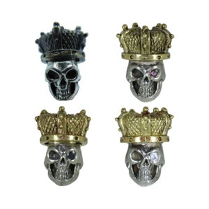 Op de afbeelding: Vier kleine schedelbedels met kronen. Drie hebben goudkleurige kronen en één heeft een donkergrijze kroon. Eén schedel heeft een rode edelsteen in de oogkas. De schedels zijn zilverkleurig.