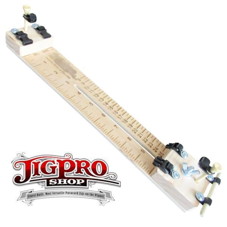 May include: A wooden jig with a ruler marked in inches and centimeters. The jig has two adjustable clamps and a small peg at one end. The jig is used for making paracord bracelets.