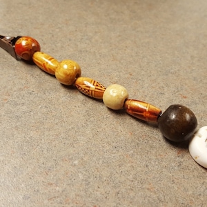 Puede incluir: Una pinza para el pelo marrón con una serie de cuentas de colores. Las cuentas son marrones, naranjas, blancas y de color marrón claro con un patrón geométrico. La última cuenta es una concha blanca.