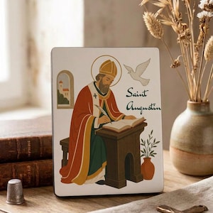 Könnte beinhalten: Eine rechteckige Holztafel mit einer farbenfrohen Illustration des heiligen Augustinus, der an einem Schreibtisch in ein Buch schreibt. Das Kunstwerk zeigt eine weiße Taube, einen goldenen Heiligenschein und den Text Saint Augustin. Die Tafel hat einen weißen Hintergrund.