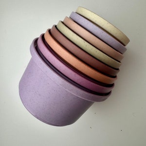 Puede incluir: Un juego de siete cuencos encajables en varios tonos de morado, rosa y beige. Los cuencos están hechos de un material moteado y están apilados en orden de tamaño, con el cuenco más grande en la parte inferior.